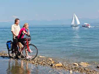 Zwei Radfahrer mit bepacktem Fahrrad am Bodenseeufer. Segelboot und Motorboot auf blauem Wasser mit Bergen im Hintergrund sichtbar.