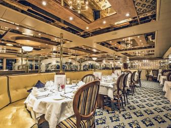 Elegantes Restaurant an Bord der MS Swiss Crown mit weißen Tischdecken, gestreiften Polsterstühlen, goldenen Sitzbänken und verspiegelter Decke.