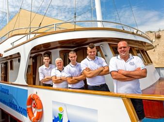 Fünf Crewmitglieder in weißen Poloshirts stehen mit verschränkten Armen an Deck der MS Amore. Blauer Himmel und historische Gebäude im Hintergrund.