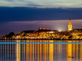 Nachtaufnahme der Friedrichshafener Skyline am Bodensee mit beleuchteten Gebäuden und Kirchturm, die sich im ruhigen Wasser spiegeln.