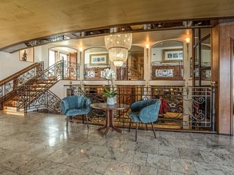 Elegante Lobby der MS Swiss Crown mit Marmorböden, verzierter Treppe, Kristallleuchter, zwei blauen Sesseln und dekorativen Geländern.