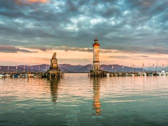 Lindauer Hafeneinfahrt am Bodensee mit Leuchtturm und Löwenstatue in der Abenddämmerung. Boote liegen im ruhigen Wasser unter dramatischem Wolkenhimmel.