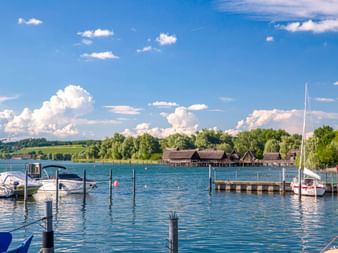 Hafen in Unteruhldingen am Bodensee mit Booten, Holzsteg und historischen Pfahlbauten im Hintergrund unter blauem Himmel mit Wolken.