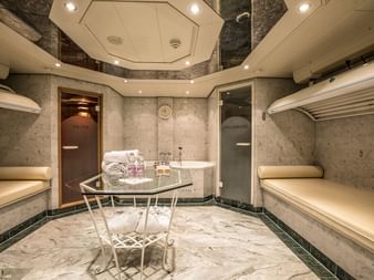 Eleganter Wellnessraum auf der MS Swiss Crown mit Marmorwänden, gepolsterten Bänken, Glassaunatür und kleinem Tisch mit Wellnessprodukten.
