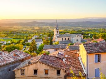 Luftaufnahme von Bonnieux mit Terrakottadächern, Kirchturm und sanften Hügeln der Provence bei Sonnenuntergang. Grünes Tal und Berge im Hintergrund.