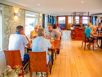 Heller Speisesaal an Bord der MS Lena Maria mit Holzboden und Tischen. Gäste sitzen an Tischen und genießen Mahlzeiten nahe großer Fenster.