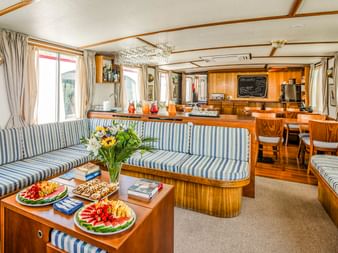 Innenraum des MS Caprice Salons mit blau-weiß gestreiften Polstersitzen, Holzmöbeln, Obsttellern auf dem Couchtisch und Küchenbereich im Hintergrund.