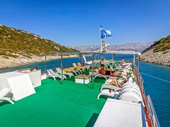 Sonnendeck der MY Mystic mit grünem Boden, weißen Liegen und entspannenden Passagieren. Blaues Küstenwasser und felsige Hügel im Hintergrund.