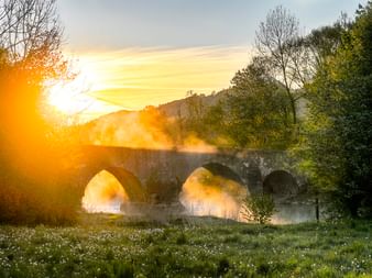 Historische Steinbrücke mit drei Bögen über die Altmühl bei Pfünz im Sonnenaufgang. Morgennebel steigt vom Wasser auf, umgeben von grünen Wiesen.