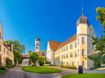Gelbes Barockschloss mit Turm in Isny im Allgäu. Grüne Rasenfläche im Vordergrund, Kirche mit Zwiebelturm dahinter, umgeben von Bäumen unter blauem Himmel.