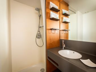 Kompaktes Badezimmer auf der MS Ave Maria mit weißer Dusche, Holzwandpaneel mit Regalen, dunkler Arbeitsplatte mit ovalem Waschbecken und Spiegel.