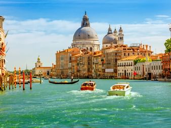 Boote auf dem Canale Grande in Venedig mit historischen Gebäuden und der Kuppelbasilika Santa Maria della Salute unter blauem Himmel.