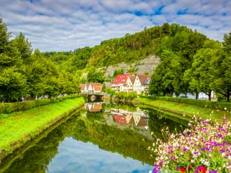Kanal mit grünen Ufern und bunten Blumen in Sulz am Neckar. Traditionelle Häuser mit roten Dächern spiegeln sich im ruhigen Wasser vor bewaldeten Hügeln.
