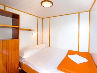 Kompakte Kabine auf der MS Planka mit Doppelbett, weißer Bettwäsche, orangem Handtuch, Holzschrank und weißen Wänden mit Holzverkleidung.