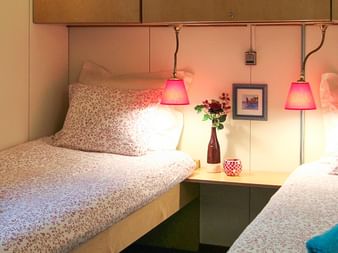 Zweibettkabine auf MS Fleur mit zwei Einzelbetten, gemusterter Bettwäsche, rosa Pendelleuchten, Nachttisch mit Blumen und türkisfarbenen und roten Handtüchern.