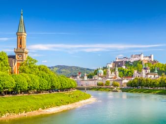 Salzach fließt durch Salzburg mit grünen Bäumen, Kirchturm und Festung Hohensalzburg auf dem Hügel unter blauem Himmel.