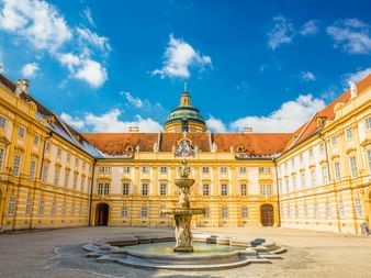 Innenhof von Stift Melk mit prächtigem Brunnen in der Mitte, umgeben von gelben Barockgebäuden mit orangen Dächern und grüner Kuppel.