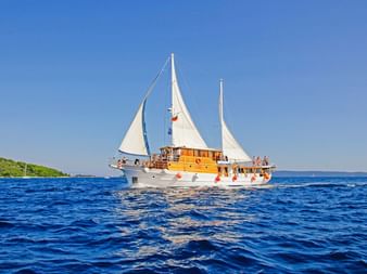 MS Mirabela, eine weiße Segelyacht mit Holz und zwei weißen Segeln, fährt auf tiefblauem Wasser unter klarem Himmel mit grüner Küste.