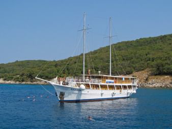 Weißes Motorsegelschiff MS Kazimir mit zwei Masten vor Anker in ruhigem blauen Wasser. Grüne Hügel im Hintergrund, Schwimmer im Wasser.
