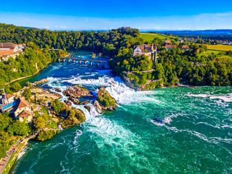 Luftaufnahme des Rheinfalls bei Schaffhausen mit türkisfarbenem Wasser, das über Felsen strömt, umgeben von grünen Wäldern, historischen Gebäuden und einer Brücke.