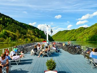 Sonnendeck der MS Normandie mit Passagieren auf Liegestühlen. Fahrräder sind an beiden Seiten geparkt. Grüne Hügel und Fluss im Hintergrund.