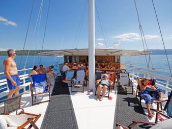Passagiere entspannen auf Sonnenliegen auf dem Oberdeck der MS Kazimir. Das Deck verfügt über einen überdachten Essbereich und Mast, blaues Wasser ist sichtbar.
