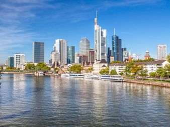 Frankfurter Skyline mit modernen Wolkenkratzern und historischen Gebäuden entlang des Mains. Boote liegen am Flussufer unter blauem Himmel.