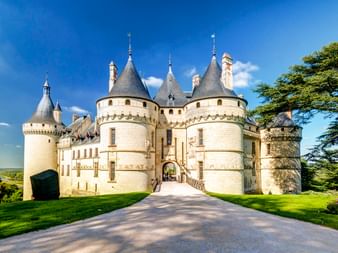 Schloss Chaumont-sur-Loire mit weißen Steinmauern, konischen Türmen und Eingangsbrücke, umgeben von grünen Rasenflächen und Bäumen unter blauem Himmel.