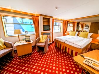 Geräumige Junior Suite auf MS Rigoletto mit Doppelbett, Sitzbereich, großen Fenstern, rot gemustertem Teppich und warmen Holzmöbeln.