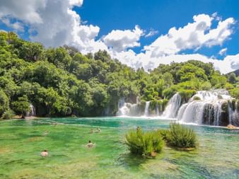 Bristane Wasserfälle mit türkisfarbenem Wasser, Schwimmer im natürlichen Becken, üppigem grünen Wald und weißen Wolken am blauen Himmel.