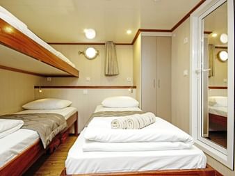 Kompakte Kabine auf der MS Aria mit Etagenbett, Einzelbett mit weißer Bettwäsche und Handtüchern, beigem Schrank, Bullauge und Holzboden.