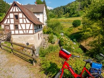 Rotes Fahrrad vor traditioneller Fachwerksmühle in Wutach, Schwarzwald. Grüne Hügel und Wald im Hintergrund sichtbar.