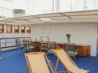 Sonnendeck der MS Azimut mit beigen Liegestühlen auf blauem Teppich, überdachtem Sitzbereich mit Holzmöbeln und weißem Sonnensegel.