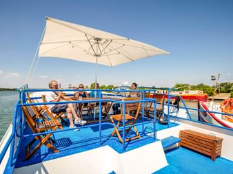 Sonnendeck des Schiffes Caprice mit blauem Geländer, weißem Sonnenschirm und Holzliegestühlen. Drei Personen entspannen im Schatten mit Flussblick.