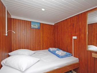 Kompakte Kabine auf der MS Kazimir mit Holzwänden, weißer Decke, Doppelbett mit weißer Bettwäsche, blauen Handtüchern, Leselampe und Spiegel.