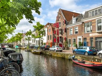 Kanal in Haarlem mit vertäuten Booten, bunten historischen Häusern, parkenden Autos am Kai und grünen Bäumen unter blauem Himmel.