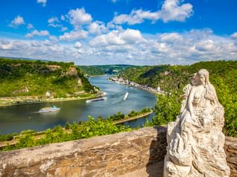 Steinstatue am Loreley-Aussichtspunkt mit Blick auf den Rhein mit Kreuzfahrtschiffen, grünen Hügeln und Sankt Goarshausen unter blauem Himmel.