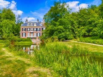 Rotes Backsteinherrenhaus mit grauem Mansardendach bei Arnheim, umgeben von grüner Parklandschaft mit Teich und Schilf unter blauem Himmel.