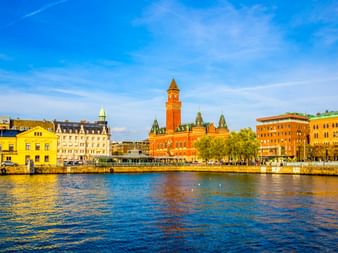 Helsingborger Uferpromenade mit dem roten Backsteinturm des Rathauses. Gelbe und beige Gebäude säumen den Hafen unter blauem Himmel.