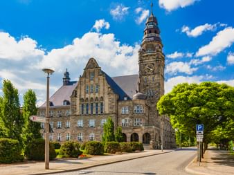 Historisches Rathaus Wittenberge mit hohem Uhrenturm unter blauem Himmel. Steingebäude mit Rundbogeneingang, umgeben von grünen Bäumen.