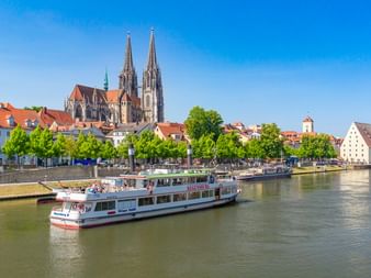 Regensburger Dom mit zwei Türmen überblickt die Donau. Ein weißes Ausflugsschiff fährt an der historischen Uferpromenade mit bunten Gebäuden vorbei.