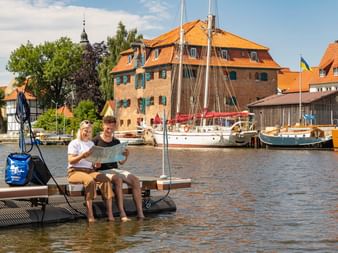 Zwei Personen sitzen auf einem schwimmenden Steg im Hafen von Glückstadt mit historischen Gebäuden, Segelbooten und orangefarbenen Dächern im Hintergrund.