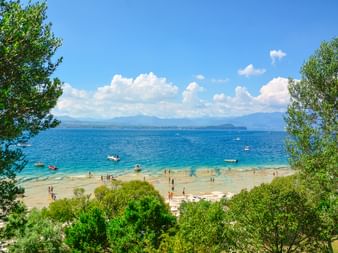 Sandstrand am Gardasee bei Sirmione mit türkisfarbenem Wasser, Booten und Menschen. Grüne Bäume rahmen die Aussicht mit Bergen in der Ferne.