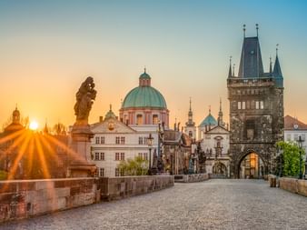 Karlsbrücke in Prag bei Sonnenaufgang mit barocken Statuen, historischen Türmen und Kirchenkuppeln. Goldenes Sonnenlicht beleuchtet die Brücke.