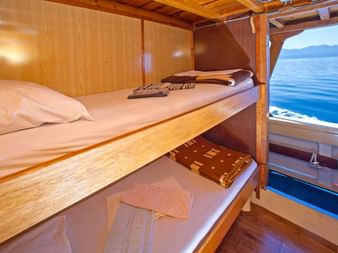 Holzkabine auf MS Aneta mit Etagenbetten, weißer Bettwäsche und großem Fenster mit Blick auf blaues Meer und ferne Berge.