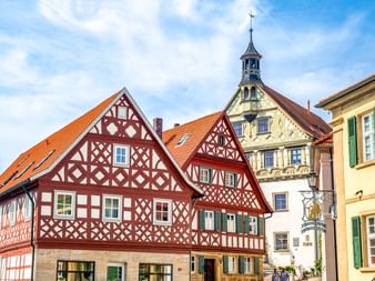 Traditionelle Fachwerkhäuser mit rot-weißen Fassaden und orangefarbenen Ziegeldächern in Burgkunstadt. Dahinter ein Kirchturm mit Uhr.