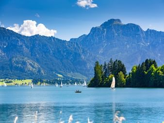 Forggensee in Bayern mit Segelbooten auf türkisfarbenem Wasser, bewaldeter Halbinsel und Tegelberg-Gebirgskette unter blauem Himmel mit Wolken.