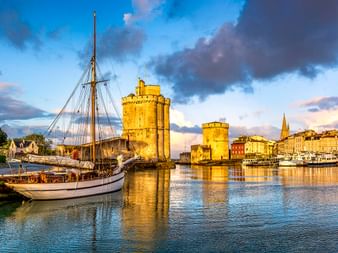 Hafen von La Rochelle mit Segelbooten, mittelalterlichen Steintürmen und Leuchtturm unter dramatischem Wolkenhimmel bei Sonnenuntergang.