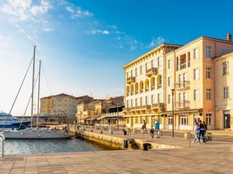 Hafenpromenade von Poreč mit Segelbooten und bunten historischen Gebäuden. Fußgänger spazieren entlang der Uferpromenade unter blauem Himmel.