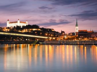 Burg Bratislava beleuchtet auf einem Hügel bei Dämmerung mit Martinsdom und Brücke. Stadtlichter spiegeln sich in der Donau.